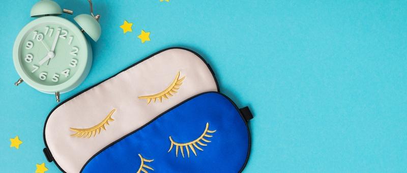 A pair of blue and pink overlapping sleep masks with eyelash detail surrounded by blue alarm clock and yellow stars, against blue background.