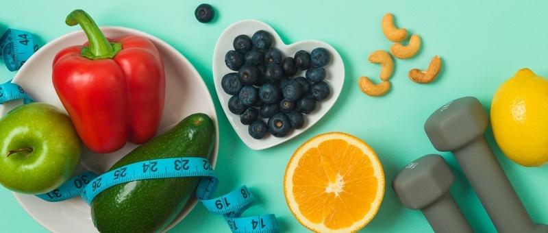 Red pepper, avocado and apple in white bowl beside white heart-shaped bowl containing blueberries - both surrounded by sliced orange, whole lemon, cashew nuts and grey dumbbells against a turquoise background with blue measuring tape.