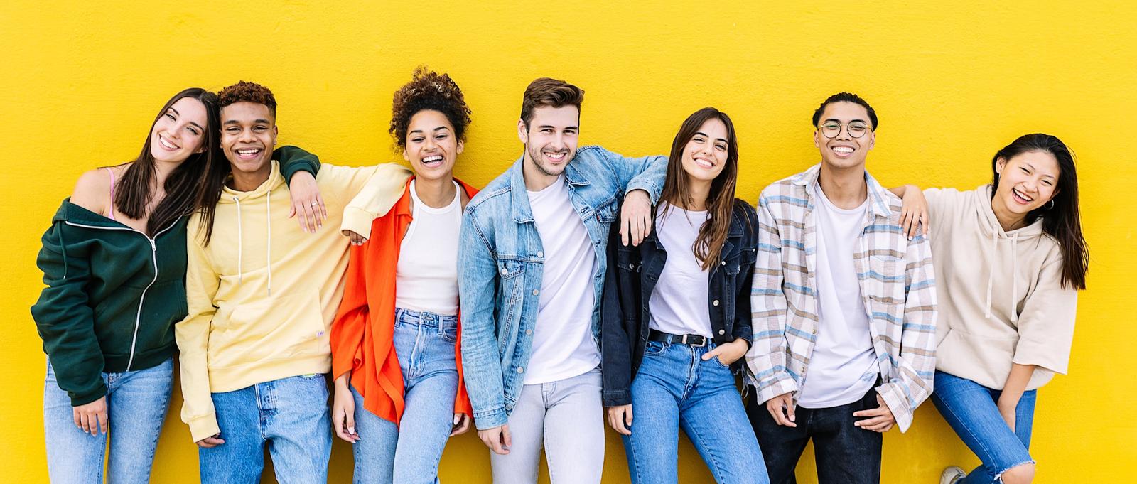 A group of students standing against a yellow wall smiling and laughing