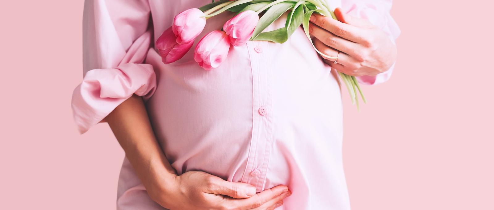 A close up of a pregnant woman's bump with her left hand holding her stomach and her right hand is resting on top whilst holding a bunch of pink tulips. The image symbolises pregnancy complications.