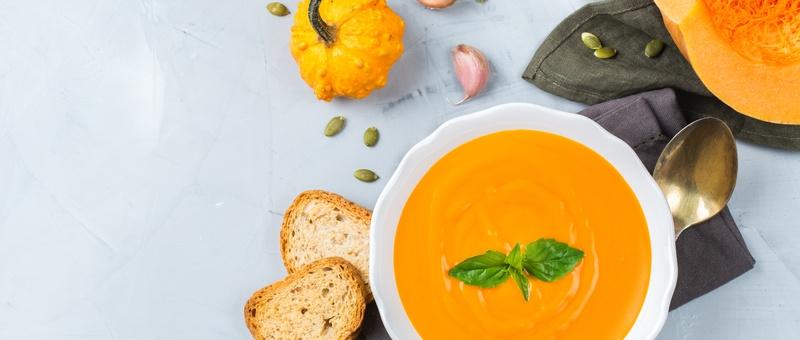 Pumpkin soup in bowl alongside bread slices, pumpkin, pumpkin seeds, metal spoon, blue napkin, and garlic bulbs against a pale grey/blue background.