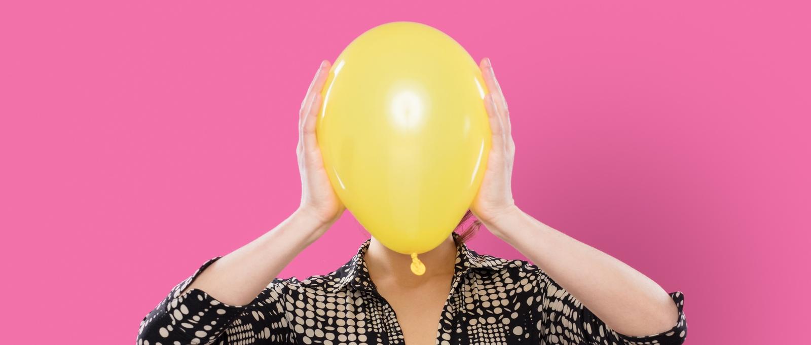 Woman against pink background holding yellow balloon over face.
