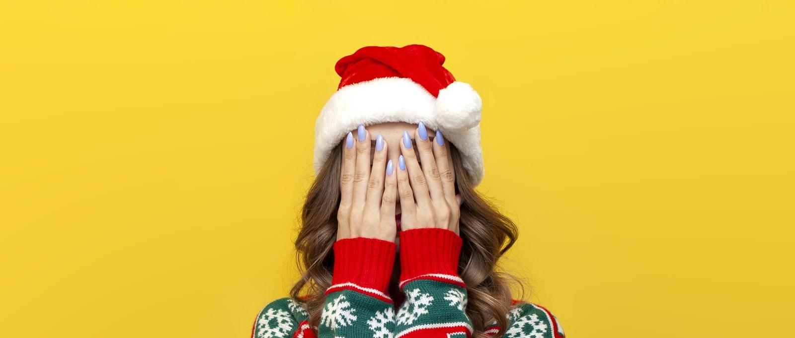 Girl in Christmas sweater and Santa Claus hat covers her face with her hand and is shy on yellow isolated background, woman hides her face in stress and depression in New Year's clothes