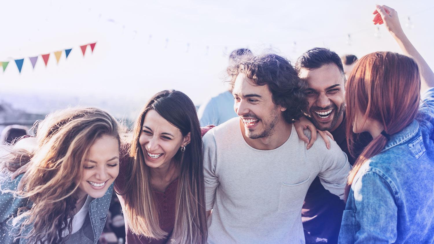 Group of friends laughing together at an outdoor celebration with colourful bunting in the background.
