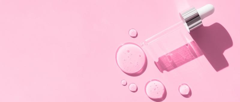 Glass dropper bottle with pink skincare serum spilled in droplets on pink background, casting subtle shadows.
