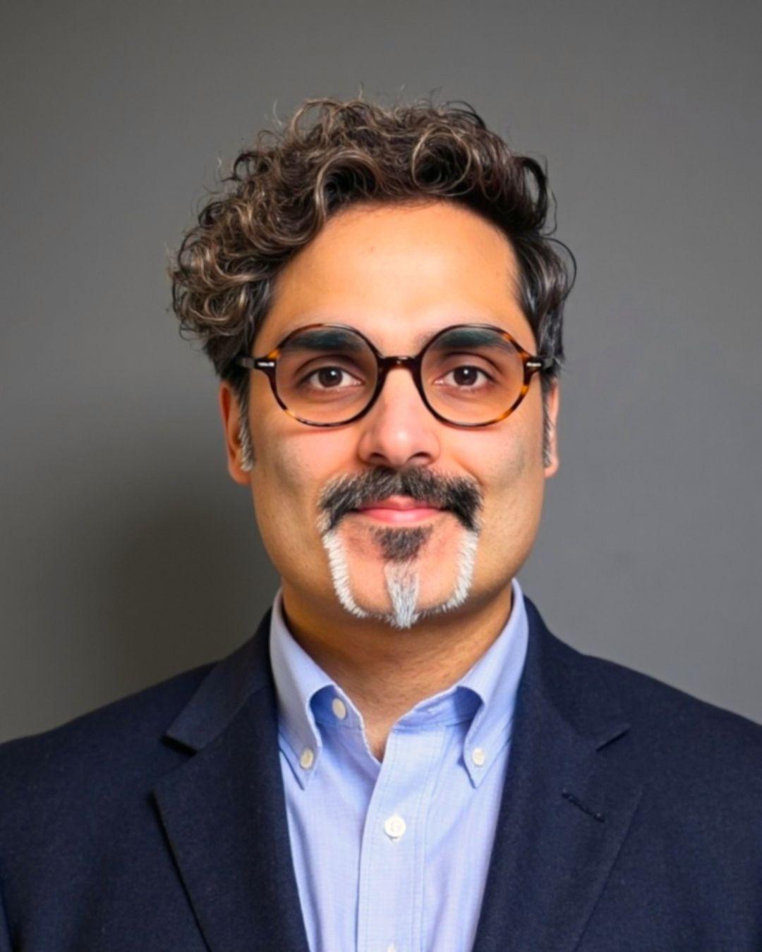 Man wearing round tortoise shell glasses with full wavy hair and trimmed beard, in a navy blue suit and pale blue shirt.