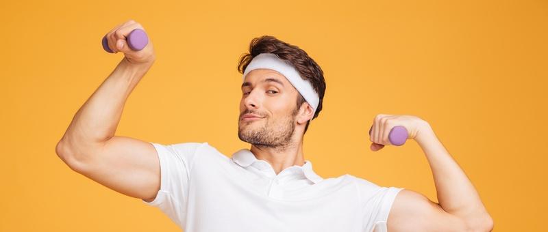 Smiling young sportsman standing and posing with dumbbells over yellow background.