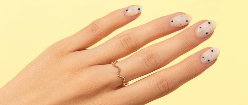 Hand with colourful polka dot nail art on nude polish wearing a delicate zigzag gold ring against yellow background.