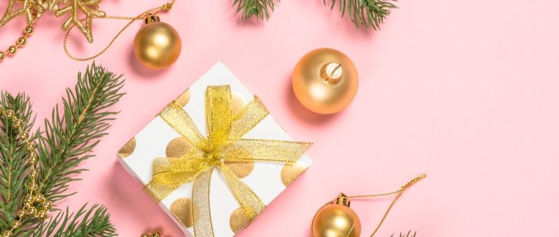 Gold Christmas tree baubles and decorations, alongside gold and white polka dot gift box with gold bow against pink background, surrounded by fir tree branches.