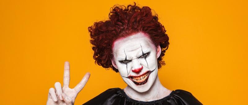 Clown with red hair in creepy black and white makeup making victory sign against yellow background.