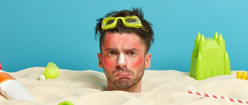 Man buried in sand with seaside toys and sunburnt face.
