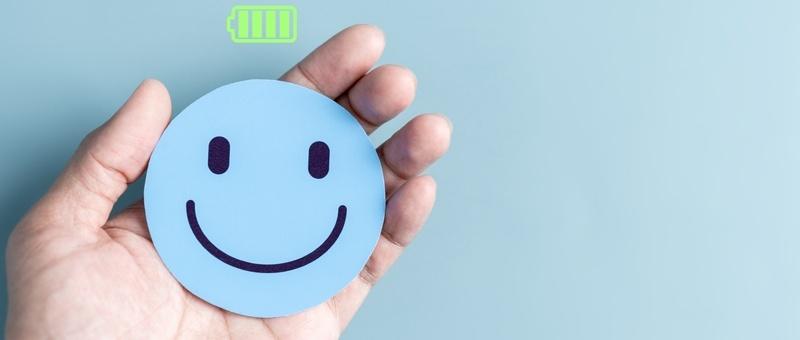 Hand holding blue cut-out smiley face beneath a green energy level battery-style bar against a blue background.