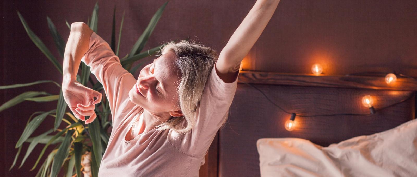 Refreshed 40s female wakes up in morning stretches seated in bed in light bedroom at home, middle-aged woman feels happy and peppy after enough sleeping, greeting new day, good morning concept