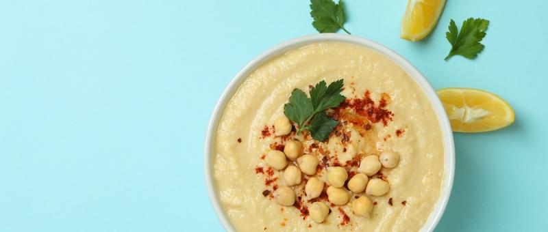 Bowl of hummus with chickpea garnish and lemon slices against a blue background.