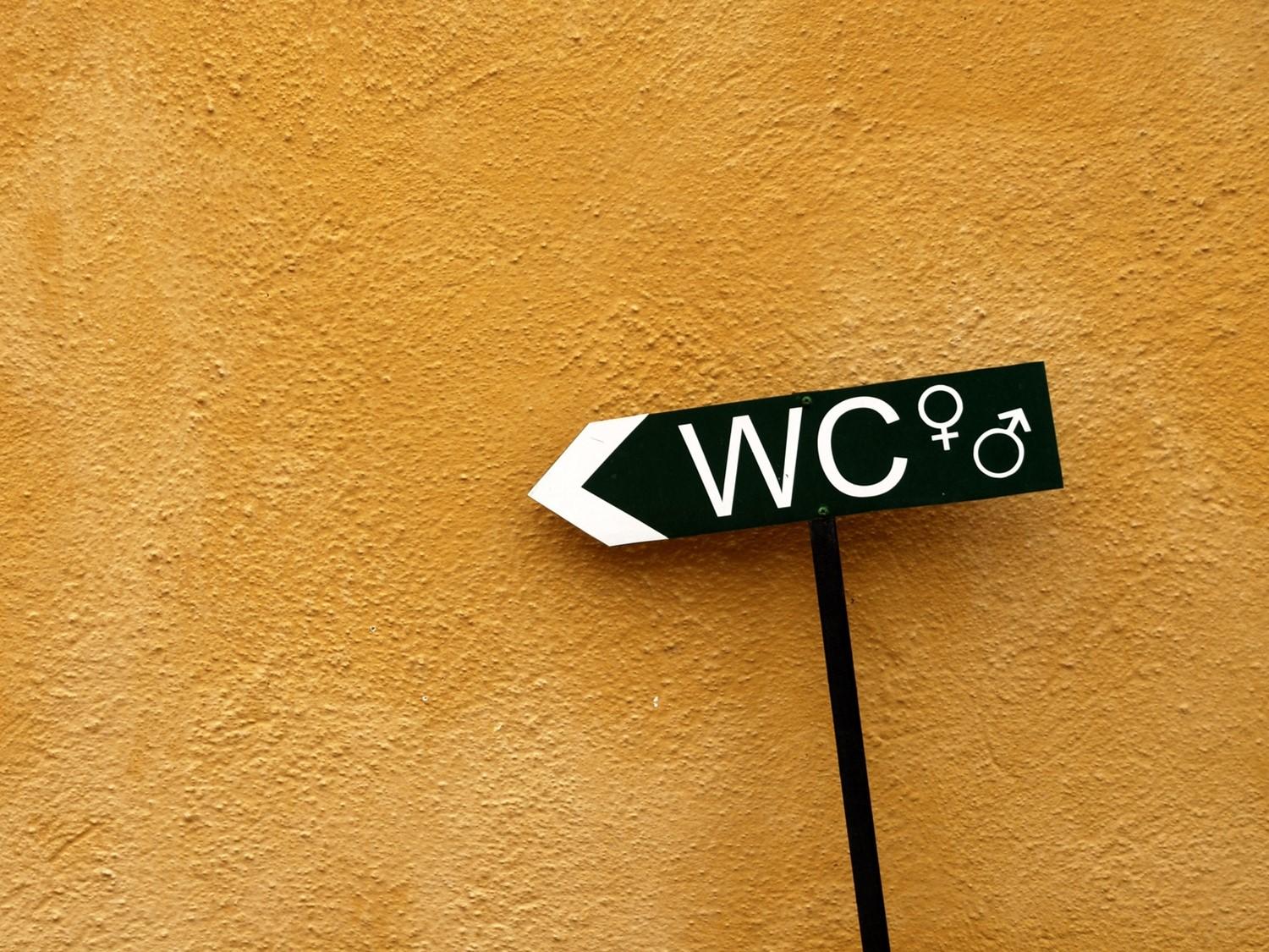 Black directional sign for toilets with male and female symbols pointing left, mounted on an orange textured wall.