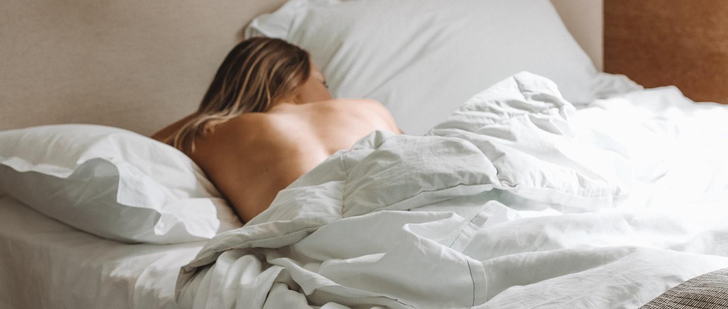 Person sleeping in bed with crisp white bedding, showing bare back against soft pillows in a neutral-toned bedroom.