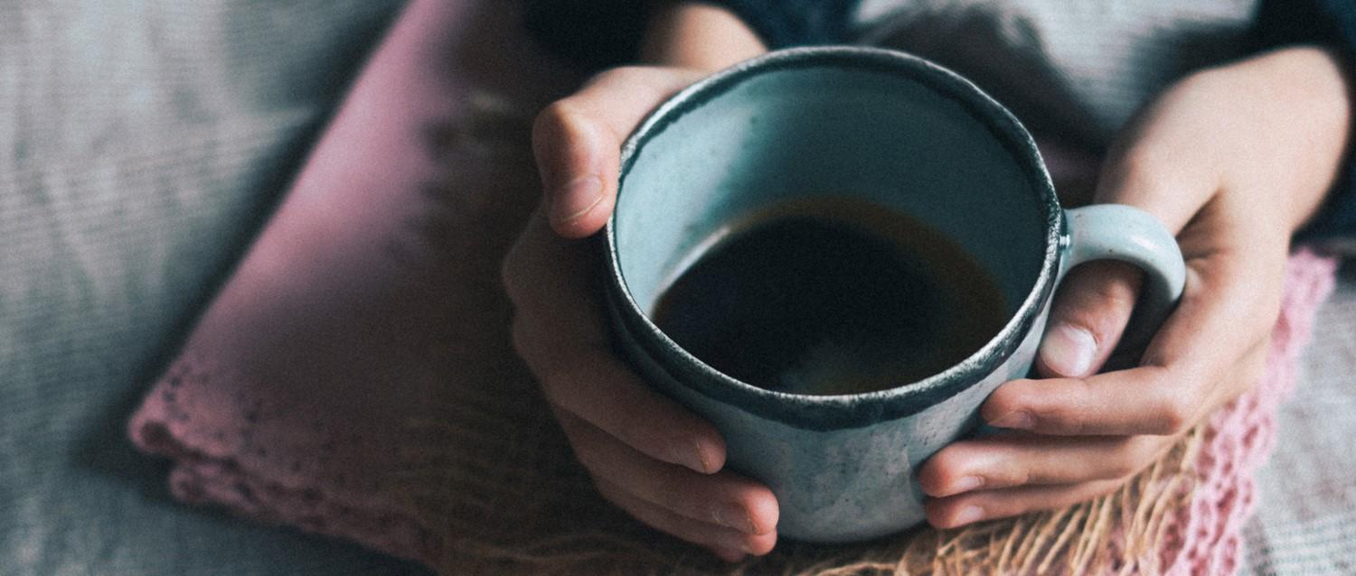 Hands holding a blue ceramic mug filled with coffee, resting on a pink textile with a cosy, intimate atmosphere.