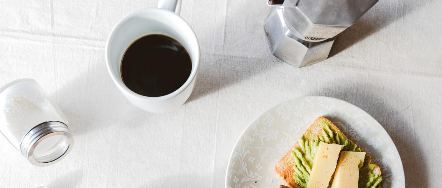 White cup of black coffee next to avocado toast with cheese on a white plate, with a moka pot and salt shaker nearby.