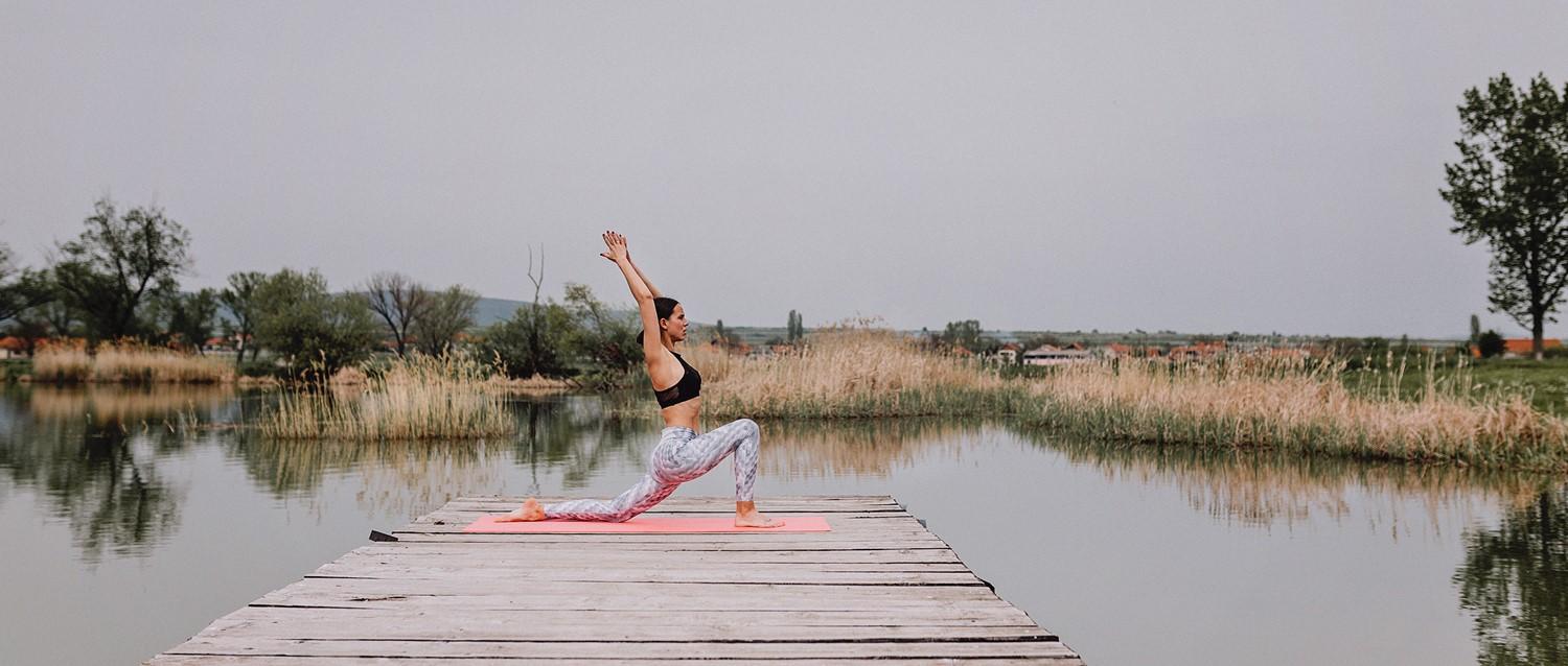 Person practicing yoga in low lunge pose on wooden dock over tranquil lake surrounded by reeds and countryside.