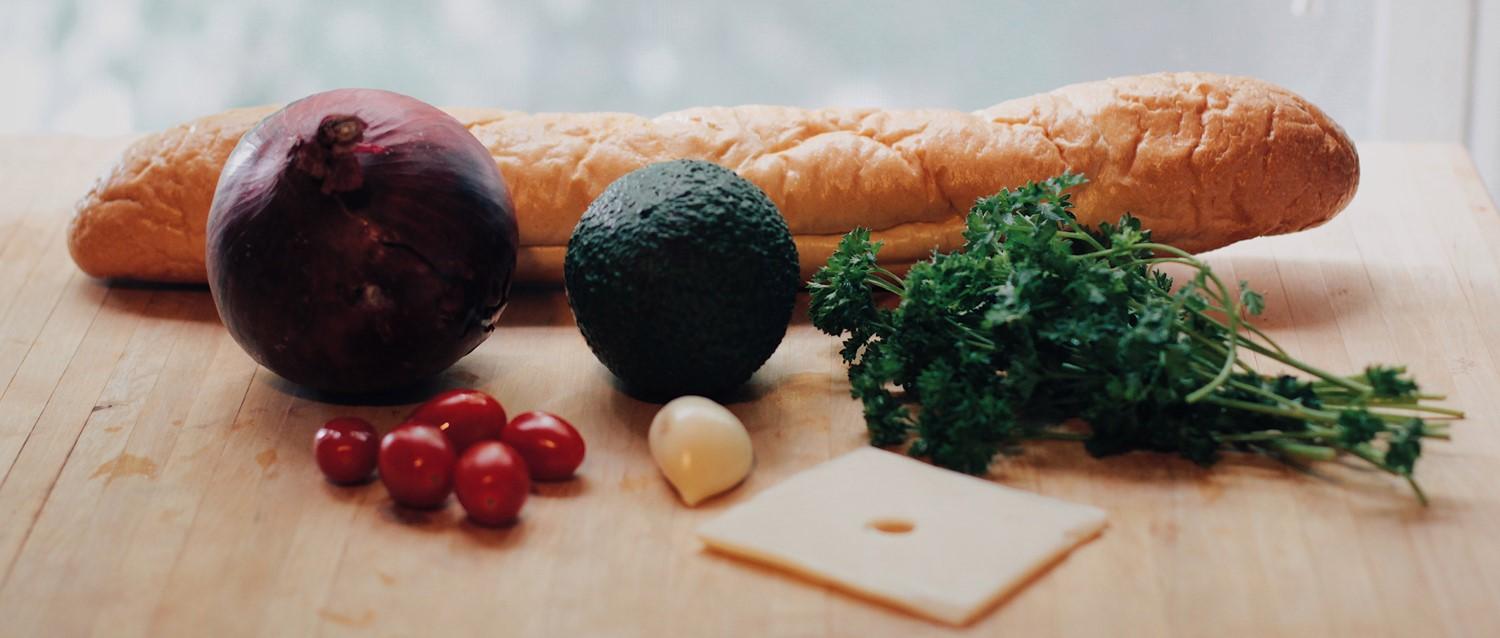 Fresh ingredients on wooden board: baguette, red onion, avocado, parsley, cherry tomatoes, garlic and cheese slice.