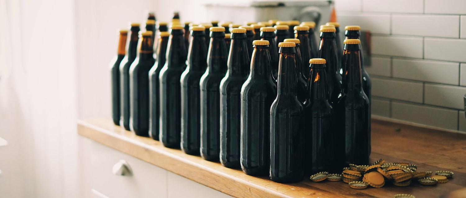 Rows of dark glass beer bottles with gold caps lined up on a wooden countertop, with bottle caps scattered nearby.
