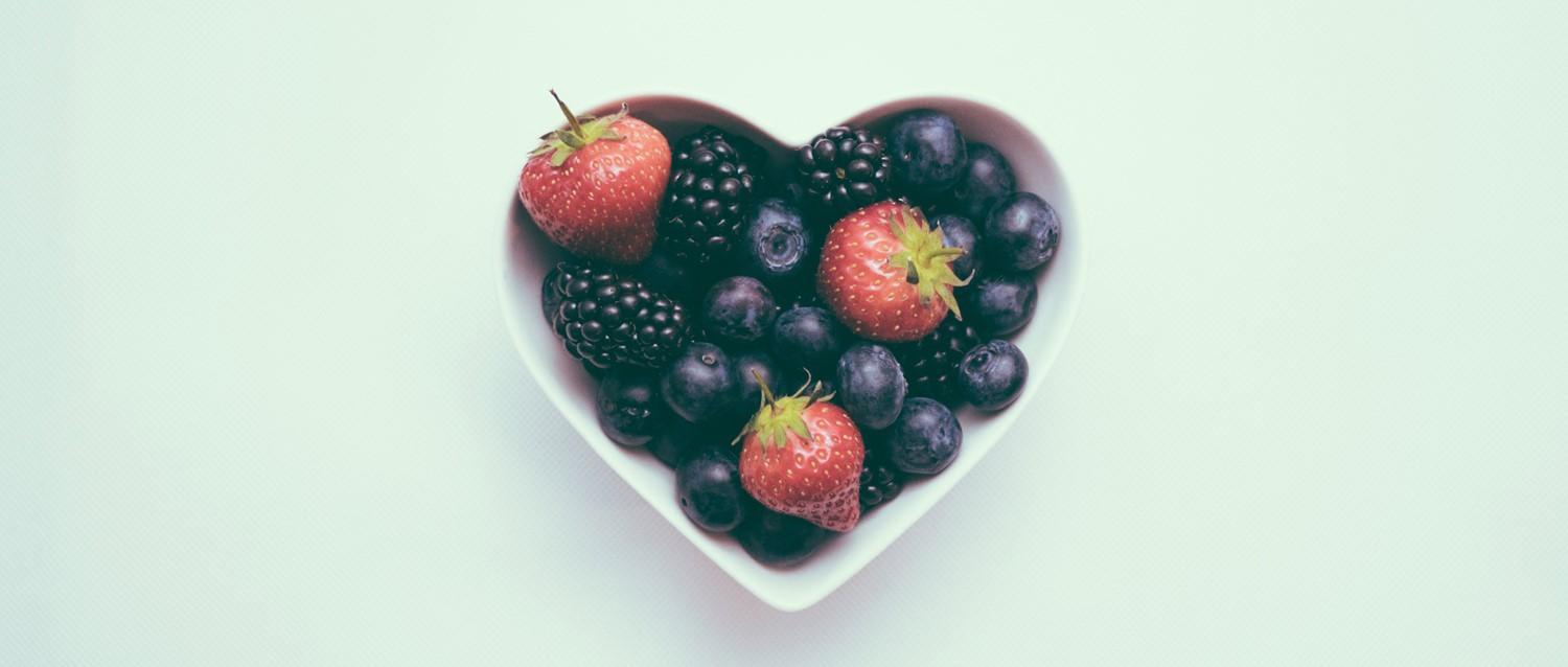 Fresh berries in heart-shaped white bowl with strawberries, blueberries and blackberries on pale mint background.