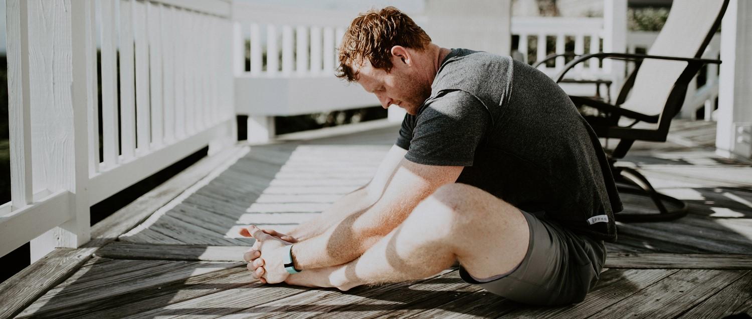 Person in grey t-shirt stretching on wooden deck with white railings, sitting in sunlight.