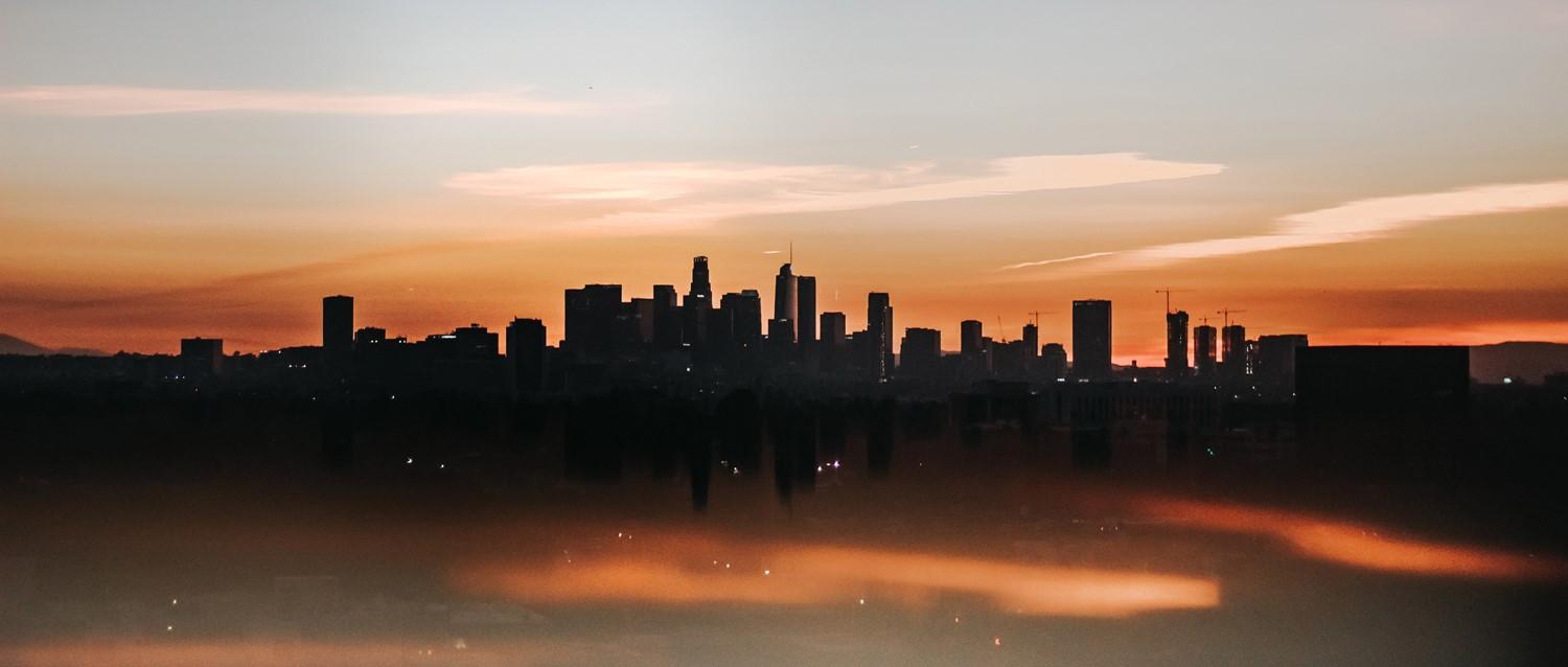 Los Angeles skyline silhouetted against a dramatic orange sunset sky, with skyscrapers reflected in water below.