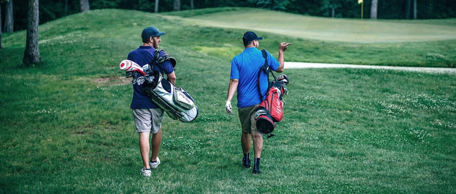 Two golfers walking across a green course carrying golf bags, one silver and one red, heading towards the next hole.