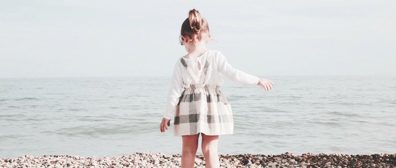 Child in white top and checked pinafore standing on pebble beach facing the calm sea on a misty day.