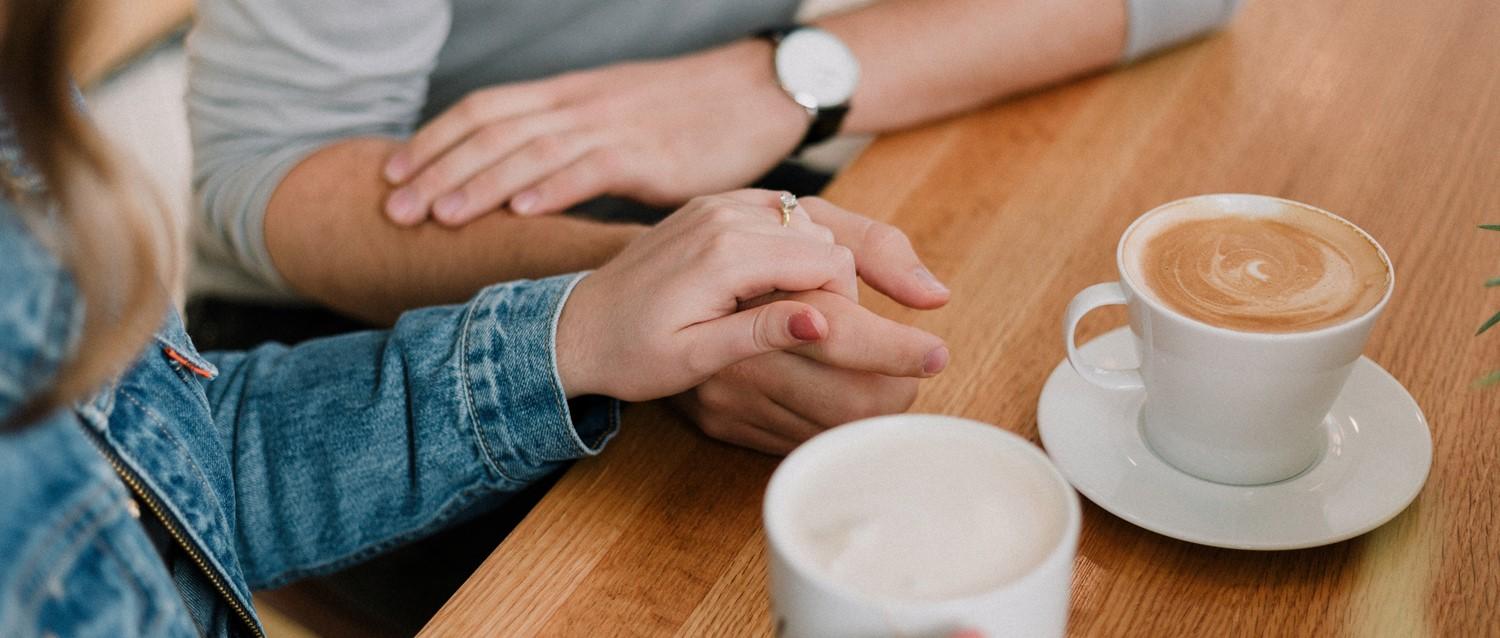 Couple holding hands across wooden table with two coffee cups, one showing latte art, intimate café moment.