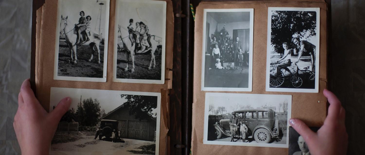 Hands holding open vintage photo album with black and white family photographs showing horses, a Christmas tree, and old car.