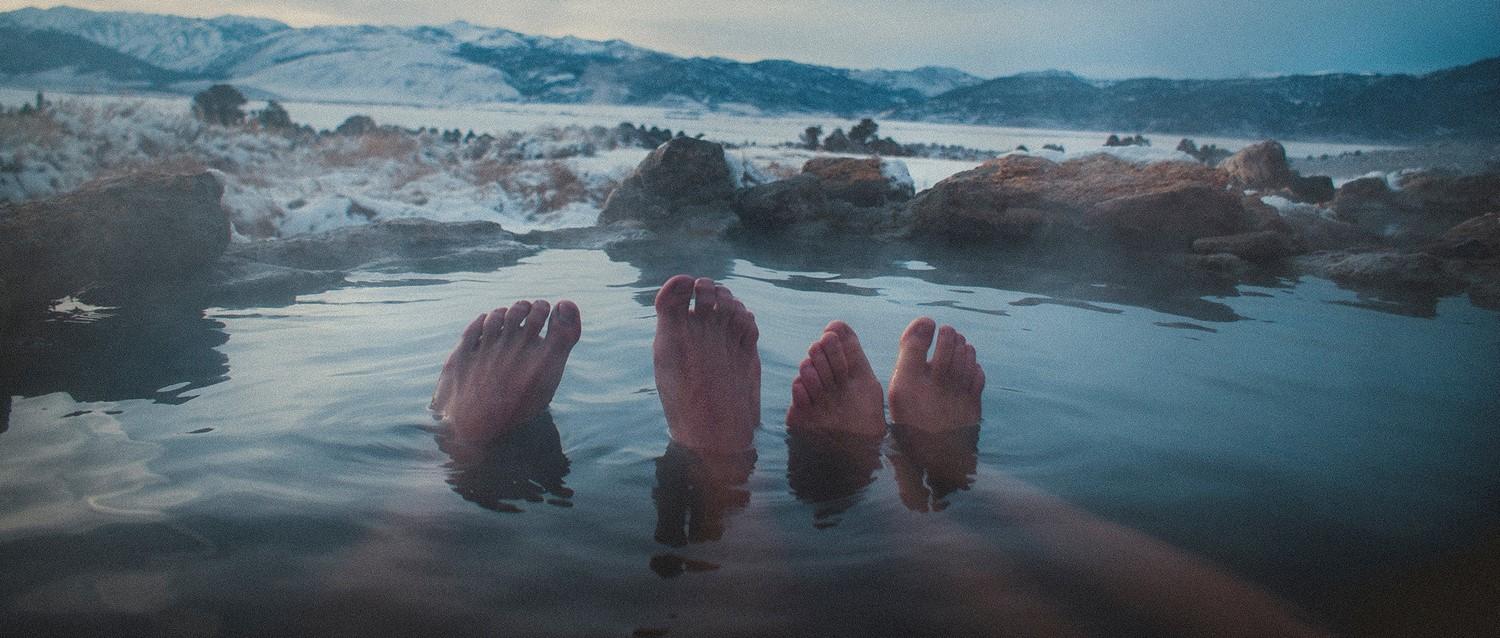 Two pairs of feet relaxing in a natural hot spring with snow-covered mountains in the background at dusk.
