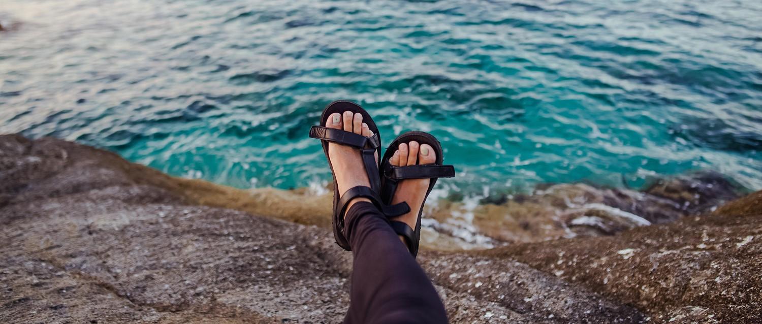 Feet in black sandals resting on rocky coastline overlooking turquoise ocean waters.