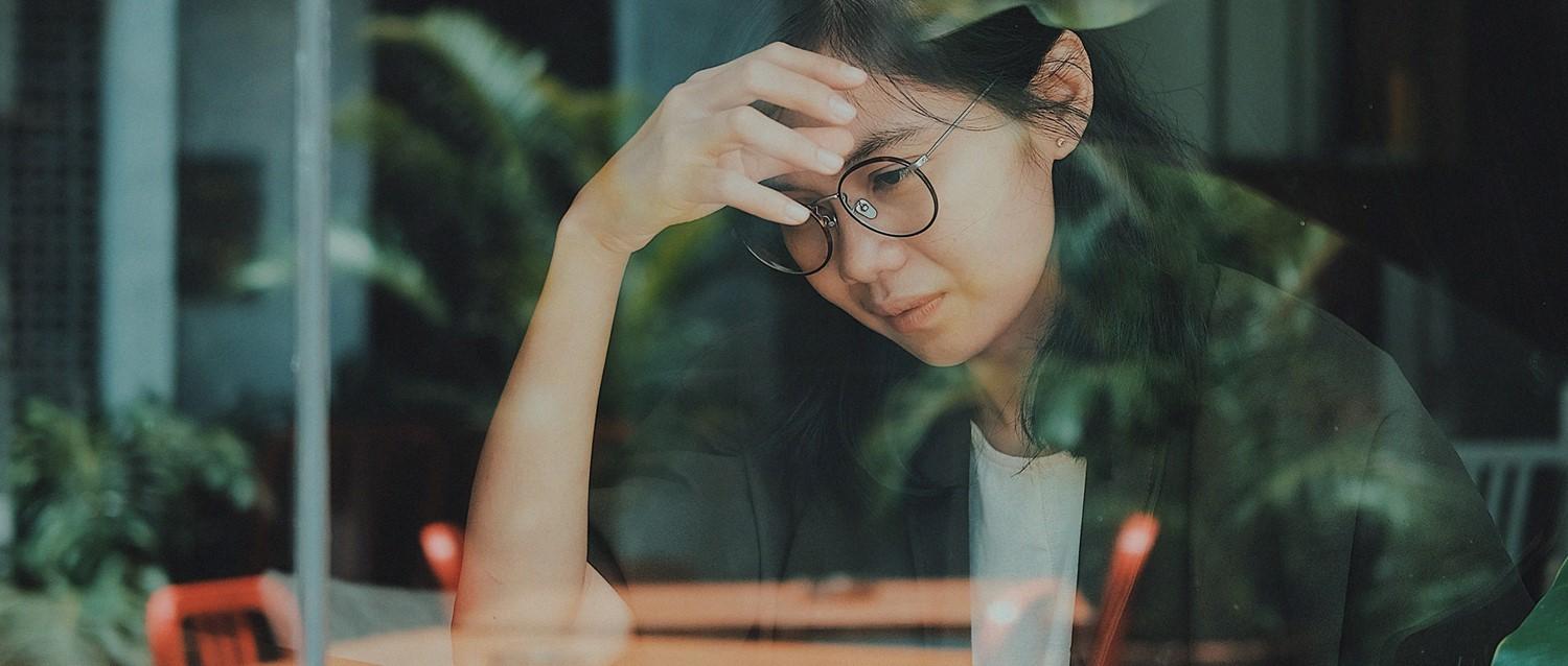 Person with glasses looking stressed, hand on forehead, sitting by window with plant reflections in muted green tones.