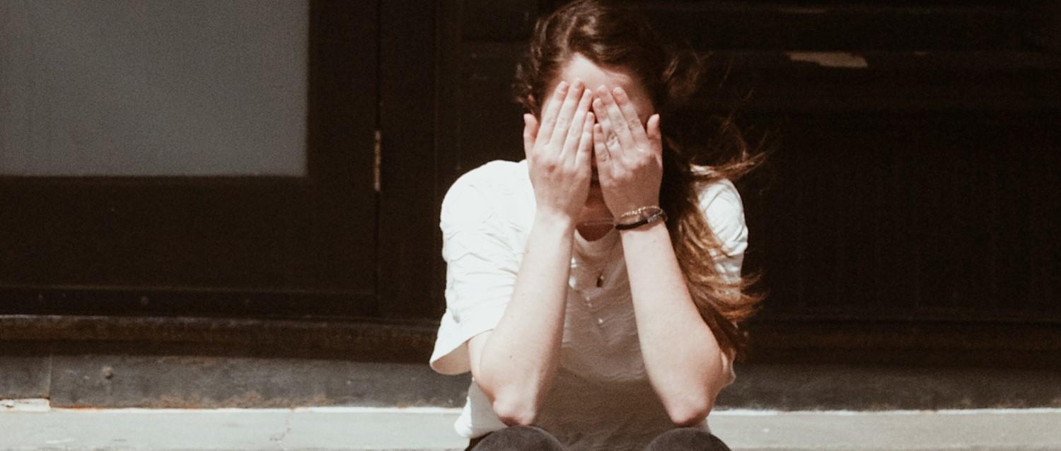 Person in white shirt covering face with hands, wearing wristwatch, sitting against dark background in vintage-toned image.