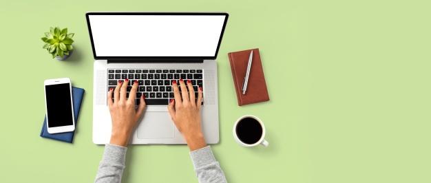 Female hands with red fingernails typing on a laptop beside phone, coffee, green succulent plant, notepad and pen, against a green background.
