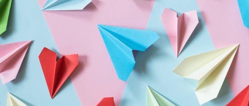 Assorted coloured paper aeroplanes and origami hearts on blue and pink background.